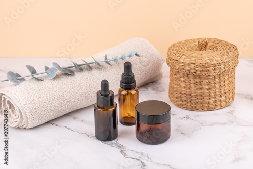 Amber glass bottles with dropper and cosmetic jar on marble surface, with rolled towel, eucalyptus branch and wicker basket. Peach background. Horizontal minimal image with copy space
