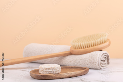 Handmade solid soap on wooden tray with wooden body brush and rolled beige towel on marble table. Peach background with soft light and relaxing atmosphere. Horizontal image with copy space