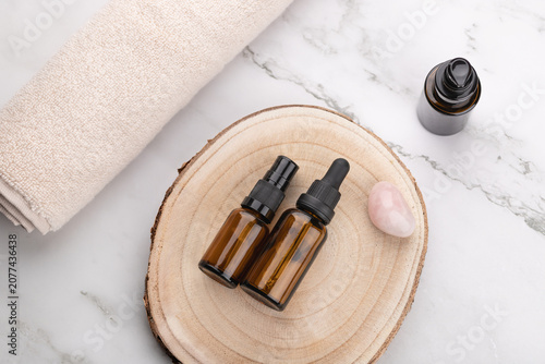 Horizontal flat lay of amber cosmetic bottles on wooden base with beige towel and rose quartz on light marble background. Minimalist style. Copy space. Concept of organic skincare and slow living