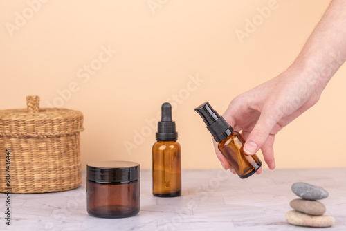 Female hand holding an amber glass bottle next to other cosmetic containers and wicker basket on marble table. Peach background with soft light. Horizontal image with copy space