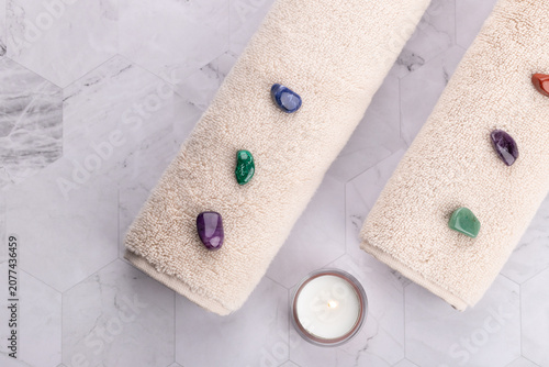 Horizontal flat lay of two rolled beige towels with colorful natural stones and small candle on stone surface. Minimal spa style with copy space. Concept of self care and mindful relaxation