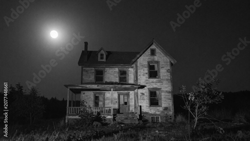 A dilapidated, two-story wooden house stands abandoned under a bright moonlit sky at night.  Haunted house in black and white.