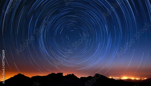 Swirling star trails above dark mountainous horizon, glowing city lights below