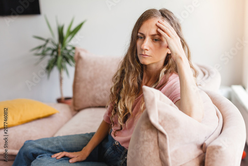Upset woman sitting on a couch at home holding her head with a hand. Stressed female feeling tired, anxious or overwhelmed by life problems. Mental health, depression and sadness concept.