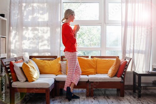 Foto Woman standing by window with cup of coffee in cozy home interior