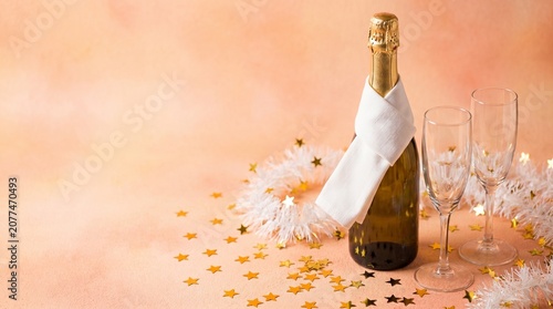 Champagne bottle with a white napkin and two empty glasses surrounded by golden star confetti on a soft peach background
