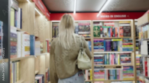 Back view of a young blonde woman walking along shelves full of books in a cozy bookstore, searching for something to read during her leisure time in a slow motion, out of focus shot