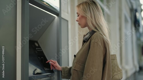 Young blonde woman using a credit card and touchscreen to withdraw money from a modern ATM machine located on a city street, completing a secure financial transaction