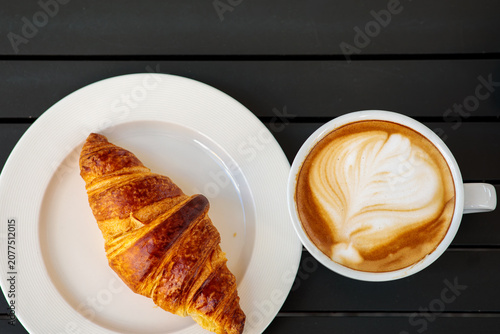 Konstfotografi A golden baked croissant rests on a white ribbed plate next to a white cup of co