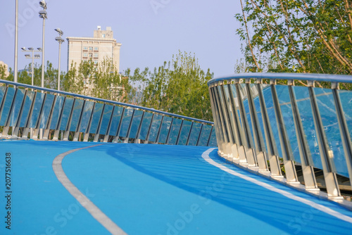 Quadro su tela A curved blue pedestrian overpass with transparent glass railings, set in an urban outdoor environment with trees and buildings