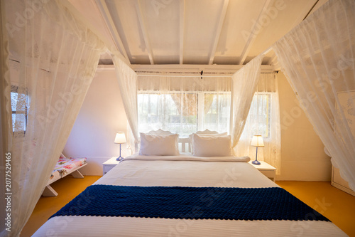 Фотография White-themed cottage house interior featuring a canopy bed with bedside lamps, a bed bench, and a sofa, showcasing a bright, elegant, and contemporary bedroom with a clean aesthetic