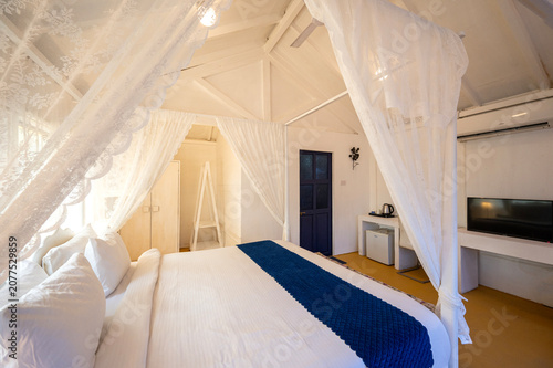 фотографія White-themed cottage house interior featuring a canopy bed with bedside lamps, a bed bench, sofa, and TV, showcasing a bright, elegant, and contemporary bedroom with a clean aesthetic