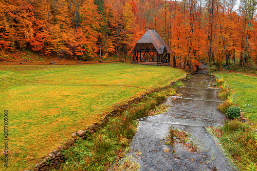 A wooden gazebo stands near a flowing stream in a park surrounded by vibrant autumn trees with orange and red foliage.
