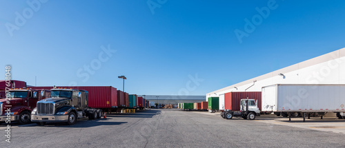 Commercial semi-trucks and shipping container trailers parked outside a large modern logistics distribution warehouse under a clear blue sky.