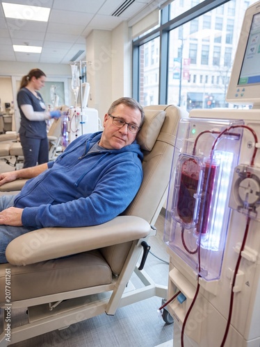 A medium shot set in a medical center for chronic kidney failure featuring a patient sitting in a reclining chair, their gaze directed toward the clear window of the dialysis machine where blood is fl