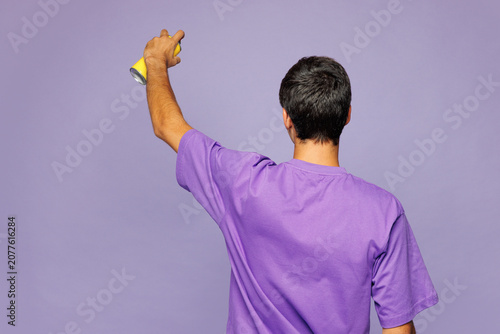 Back rear view young man he wear violet t-shirt casual clothes hold in hand spray paint bottle do graffiti stretch arm on empty wall isolated on plain pastel light purple background Lifestyle concept.