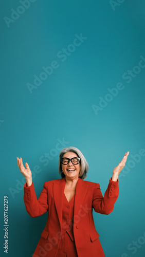 Happy senior executive woman in red pantsuit and glasses smiling, celebrating success with raised arms