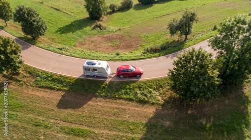 Aerial view of camper van towing red car on winding road through green countryside, road trip travel concept.