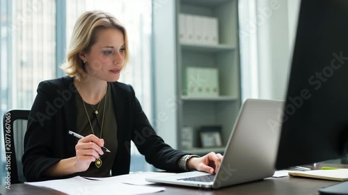 Professional businesswoman sitting at office desk, working on laptop and taking notes. Modern corporate workspace with natural daylight and organized interior.