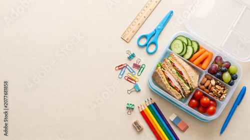 Healthy school lunch box with sandwich, fresh vegetables, fruits and nuts with colorful stationery on wooden background, top view with copy space