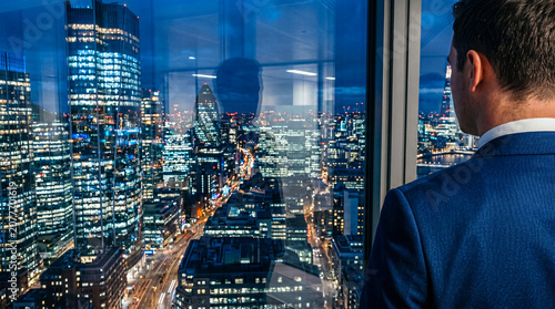 Εκτύπωση καμβά Back view of a businessman in a suit looking at a stunning illuminated cityscape from a high-rise office at night