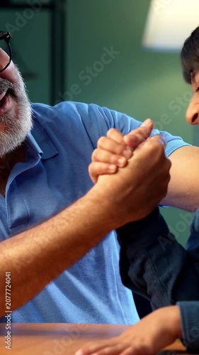 Indian grandfather and kid arm wrestling at home, both sitting together at table in modern house, elderly man and grandson enjoying playful activity, family bonding moment of two people having fun