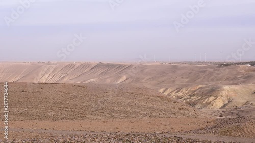 A wide view of wind turbines standing in the arid desert landscape near the historic Shobak Castle in Jordan under a soft, hazy sky.