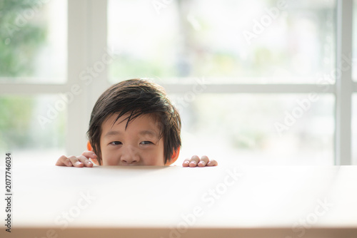 Cute Asian little boy with wet hair peeking over the edge of a wooden table indoors with copy space.
