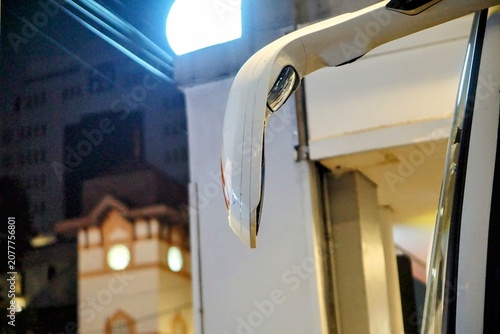 Fotografie Close-up detail of a modern giraffe-style tour bus side mirror gleaming at night with a backdrop of ancient and modern city architecture