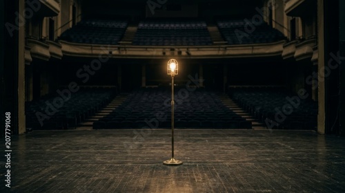 Ghost light lamp placed in the center of an empty theater stage. Single glowing bulb on a stand representing safety and tradition in performing arts. Dramatic interior lighting concept.
