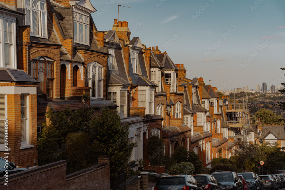 Fototapeta premium A beautiful row of classic Victorian brick terrace houses lining a steep residential street, captured during the warm golden hour sunset with parked cars reflecting the bright evening sunlight.
