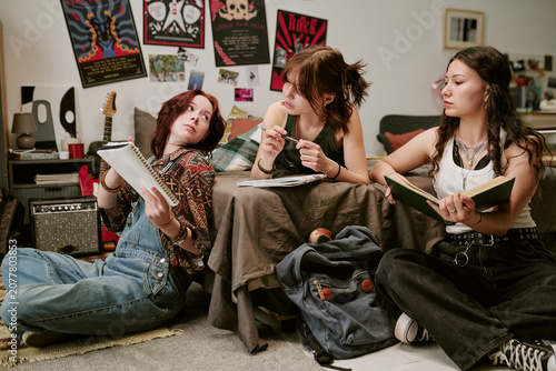 Photos Teenage girls studying together in bedroom, writing notes and reading book while discussing homework, conveying collaborative learning