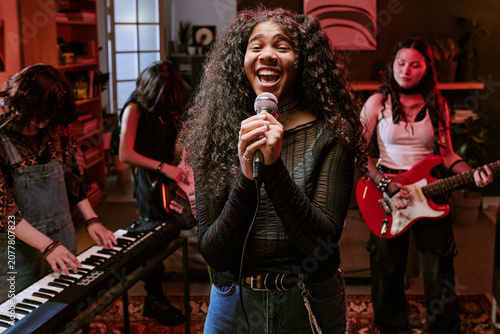 Portrait of Black teenage girl singing into microphone while teen band playing keyboard, bass, guitar in rehearsal, representing youth music culture. Ideal for music education, teen talent promotion