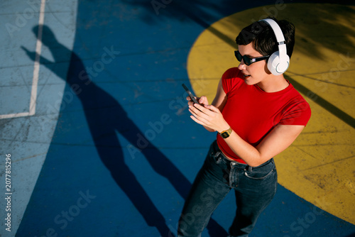 Young Caucasian woman wearing wireless headphones and sunglasses while using her smartphone in a colorful urban outdoor setting