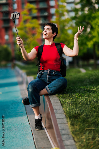 Smiling Caucasian female content creator recording a video with a smartphone mounted on a selfie stick while sitting outdoors in a city park