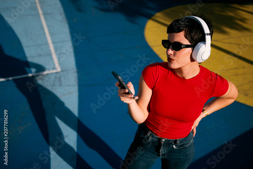 Young Caucasian woman wearing wireless headphones and sunglasses while checking her smartphone in a colorful urban outdoor setting