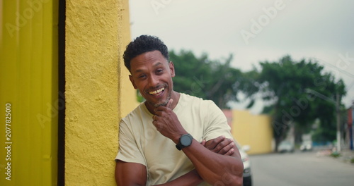 Tableau sur toile Hispanic Afro-Latin man leaning against yellow wall, smiling warmly, hand on chi