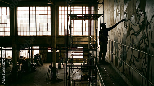 An artist creates graffiti on a wall in an abandoned industrial building with scaffolding.