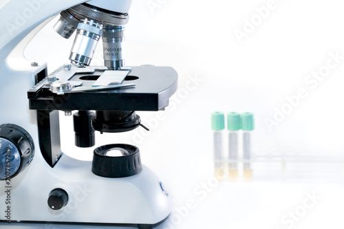 Optical microscope with sample slide and blurred laboratory test tubes on white background, medical research concept with copy space.