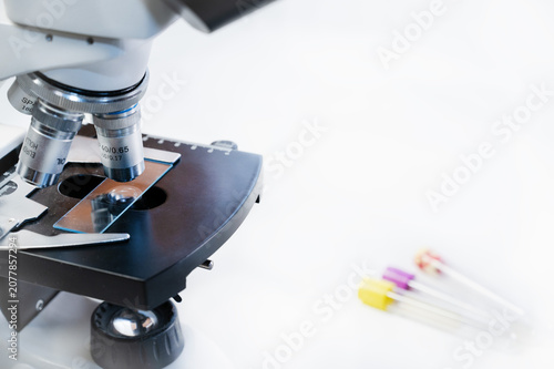 Close-up of optical microscope with glass slide and blood test tubes on white background in laboratory, copy space