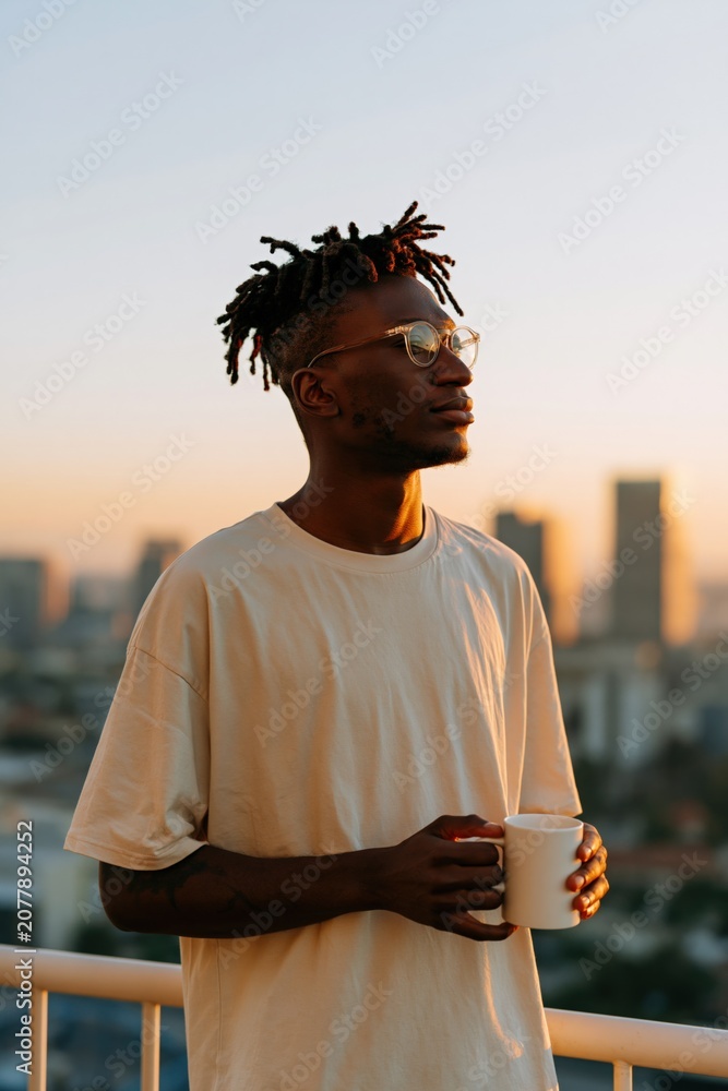 Fototapeta premium Thoughtful man with glasses holding a mug on balcony at golden hour for urban poster and lifestyle background