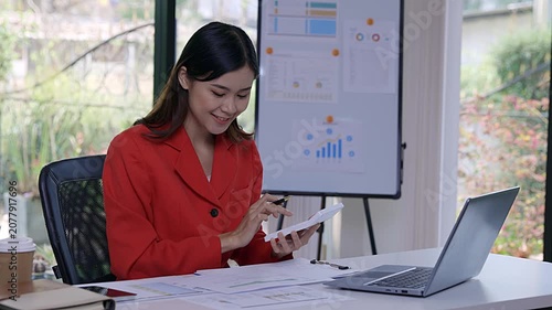 Successful Asian businesswoman in red suit using calculator to analyze financial data chart on desk at office.