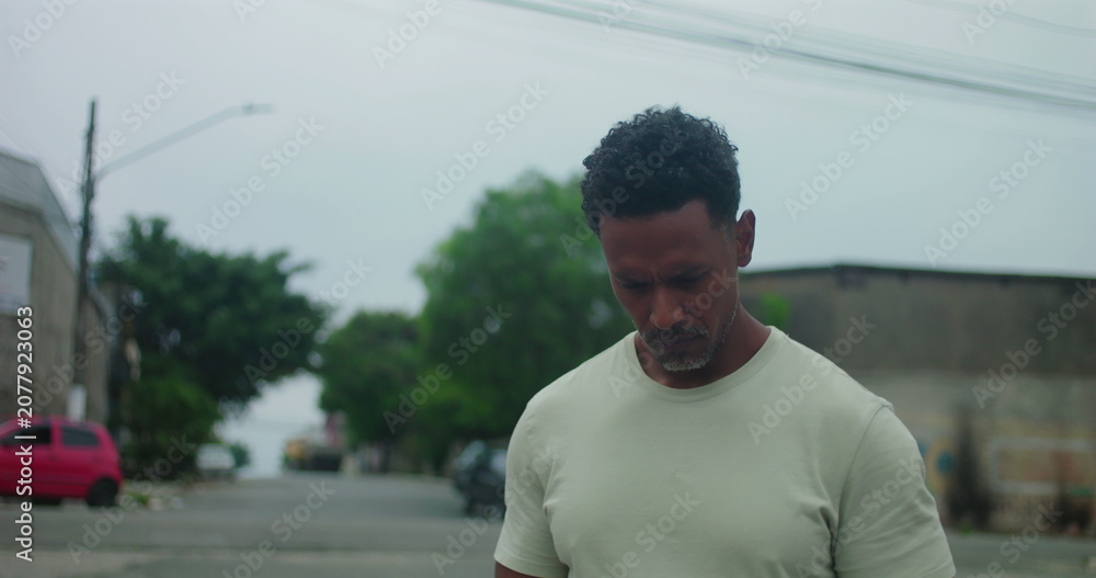 Fototapeta premium Man of African descent walking on urban street with serious expression, looking into distance, deep in thought, focused and introspective, muted cityscape, quiet determination