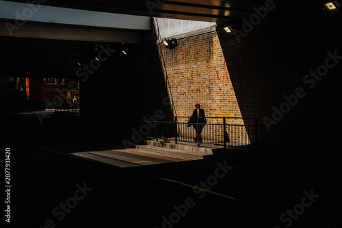 Konstfotografi Dramatic high-contrast shot of a lone pedestrian walking along a brick wall illuminated by a sharp beam of bright sunlight cutting through a dark concrete underpass tunnel