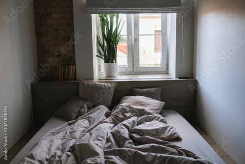 A cozy bedroom interior with a messy bed and a large window. Cozy unmade bed with a crumpled grey duvet under a bright window in soft morning light.