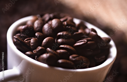 Roasted coffee beans overflowing from a white ceramic cup. A cozy scene that invites the preparation and enjoyment of coffee.