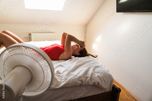 Exhausted woman cooling down near electric fan on bed during summer heatwave