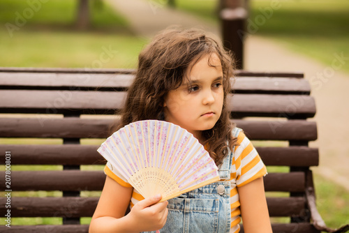 Foto Cute little girl kid child is sitting on a park bench, fanning herself with a decorative fan to cool off