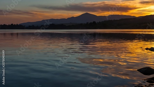 Stunning golden sunset over calm lake water with distant mountain silhouettes and warm orange glowing atmospheric light