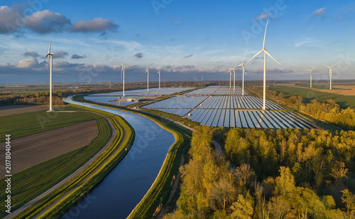Aerial view of a solar farm and wind turbines situated next to a canal in a rural landscape with green fields and trees in Groningen, Netherlands.